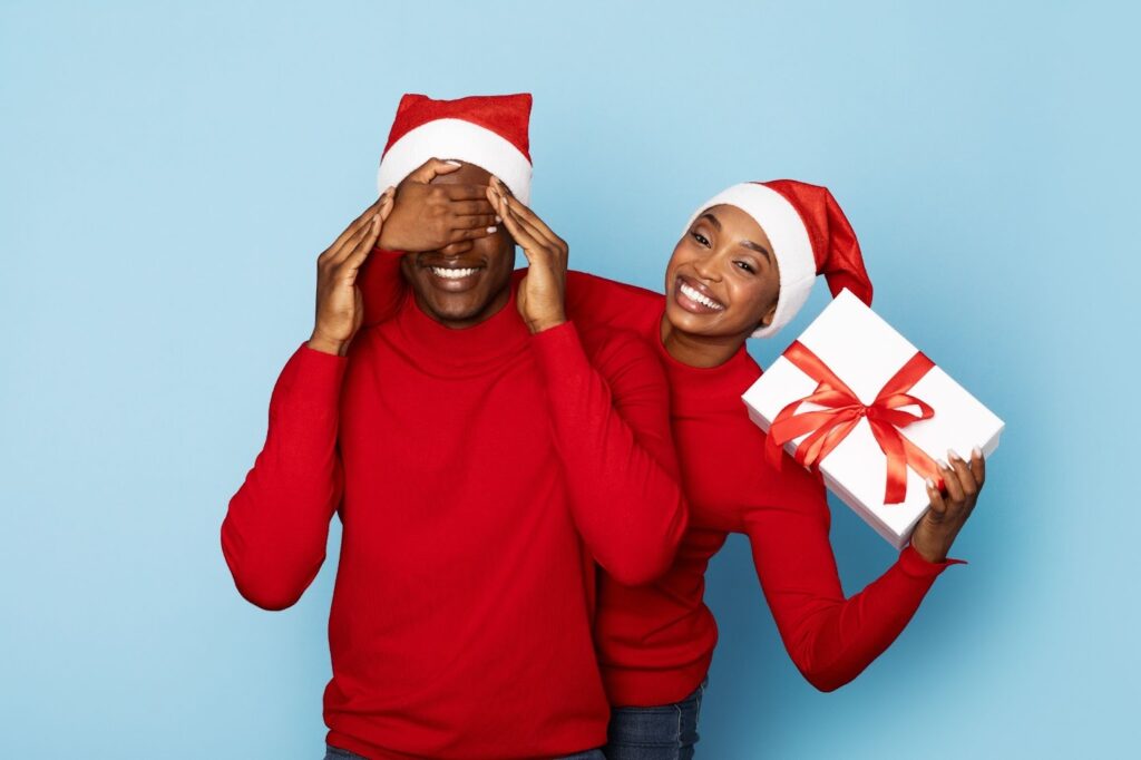 fun couple hiding a gift wearing red sweaters and a Santa hat