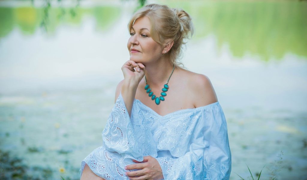 Older woman thinking by lake in a blue top